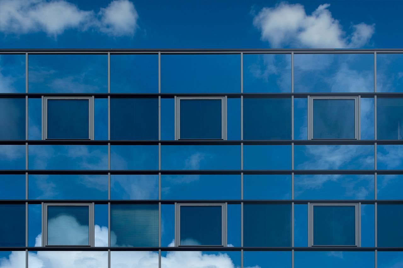 hero-img-02 Facade of a modern glass building reflecting the blue sky and clouds.